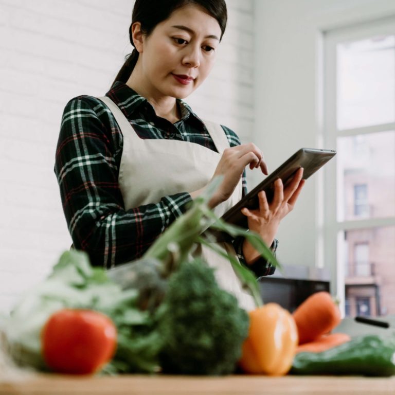 Tablier Japonais Femme portant un tablier japonais en cuisine consultante une tablette, entourée de légumes frais.