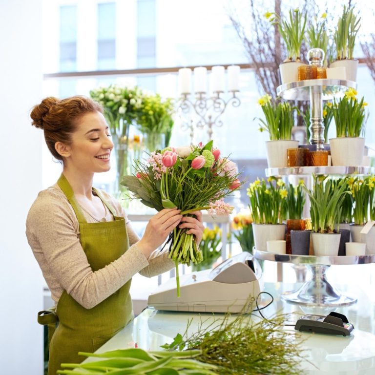 Tablier de Fleuriste en Jardinerie Femme souriante préparant un bouquet de fleurs avec un tablier vert clair dans une boutique lumineuse.