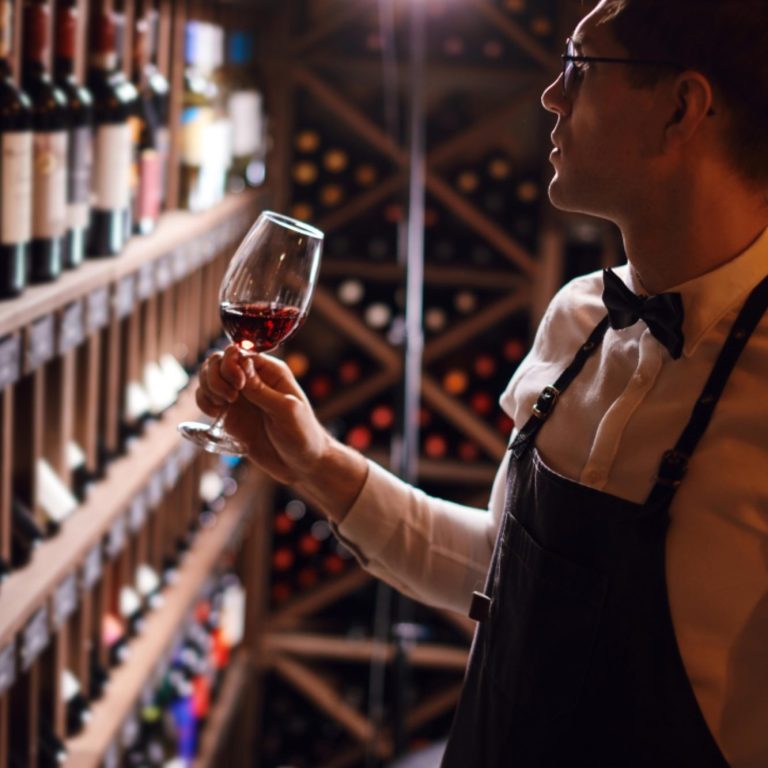 Les Tabliers de Sommelier Un sommelier examine un verre de vin rouge devant une étagère à vins.